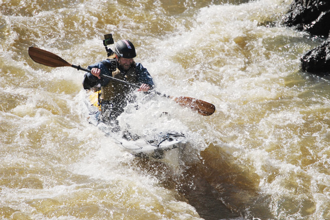 A kayak angler wearing waterproof fishing gear paddles into a big rapid.